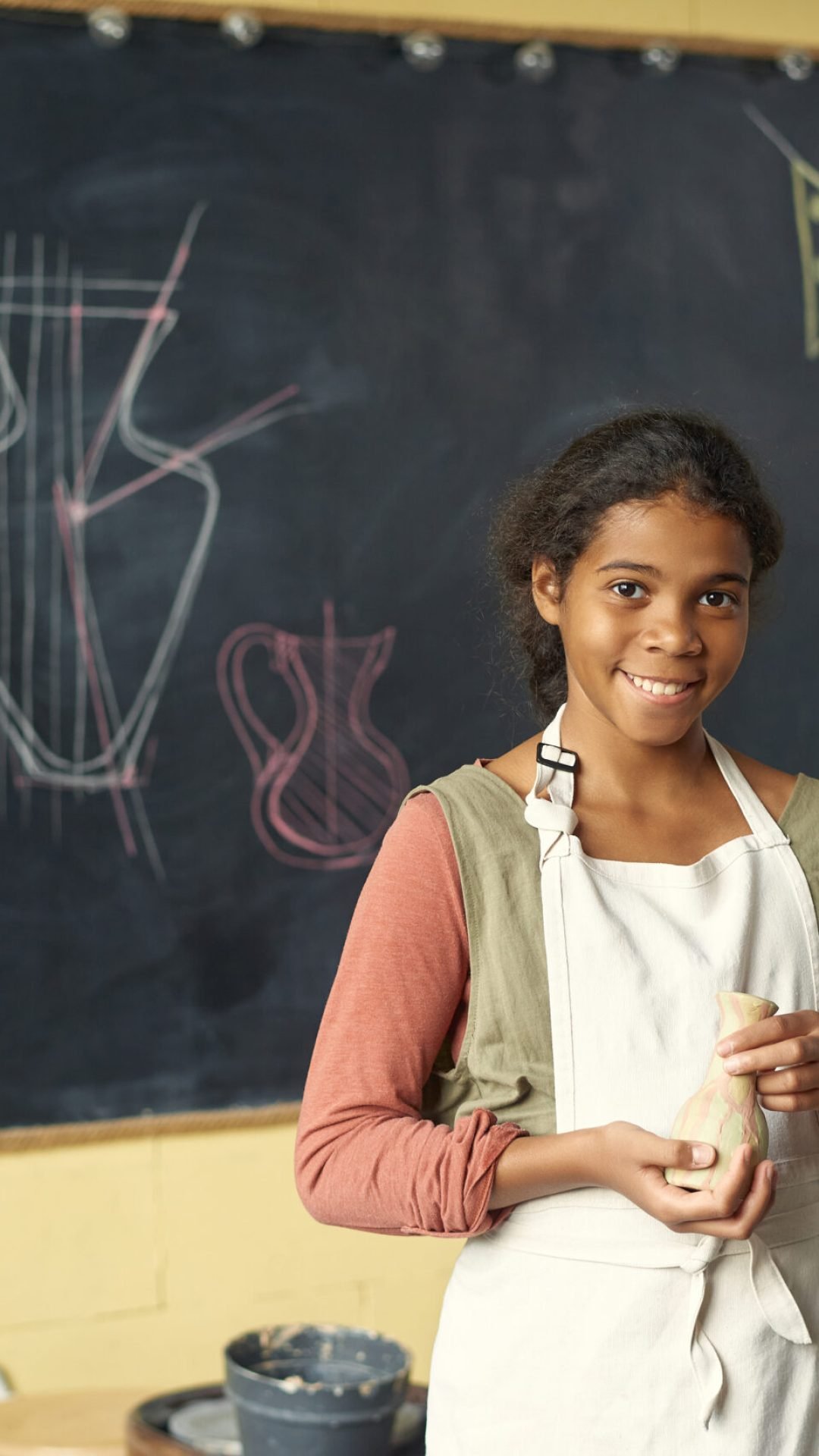 Happy cute schoolgirl with handmade clay pot standing by blackboard with sketches of earthenware in classroom